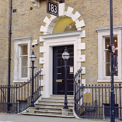 The image features a large, old-fashioned building with a black door and white trim. Above the door is a sign that reads "18." The building has a yellow archway and a balcony on the second floor. There are two street lamps positioned in front of the building, one closer to the left side and the other nearer to the right side.