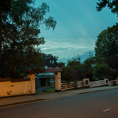 The image depicts a quiet street at dusk, with trees and buildings lining the road. There are several cars parked along the side of the street, and a few people can be seen walking or standing near them. A bench is also visible on the sidewalk, providing a place for pedestrians to sit and relax.