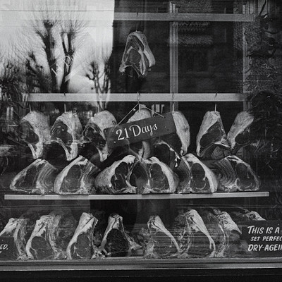 The image is a black and white photograph of a store display featuring a large number of hanging raw meat products. There are at least 13 pieces of meat visible in the display, with some hanging higher than others. The arrangement creates an eye-catching presentation for customers visiting the store.