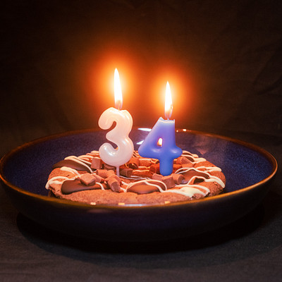 The image features a blue bowl placed on a black table, filled with chocolate candies and two lit birthday candles. The candles are positioned in the middle of the bowl, creating an eye-catching centerpiece for the celebration. The arrangement suggests that this is likely a 34th birthday party, as indicated by the number of candles on the cake.