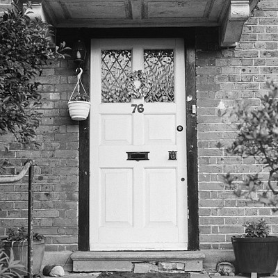 The image features a white door with a black mail slot, located on the side of a brick house. Above the door is a hanging basket and a number 86 on it. There are two potted plants in front of the door, one to the left and another to the right.