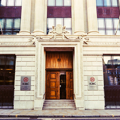 The image features a large, white building with an ornate entrance. Above the doorway, there is a sign that reads "ibc." The building appears to be a bank or financial institution, as evidenced by the presence of multiple signs on its exterior. In addition to the main entrance, there are two other doors visible in the scene.