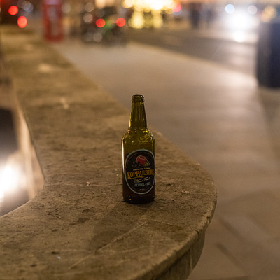 The image features a bottle of beer sitting on top of a stone ledge or counter. The bottle is placed in the center of the scene, and it appears to be an old-fashioned style beer bottle. In the background, there are several cars parked along the street, with some closer to the foreground and others further away.