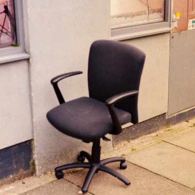 The image features a black office chair sitting on the sidewalk outside of a building. It is positioned next to a wall, and there are two windows visible in the background. The chair appears to be empty and ready for someone to sit down and use it.