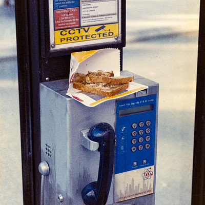 The image is a black and white photo of an old payphone with a piece of food, specifically a hot dog, sitting on top of it. The phone appears to be outdated, possibly from the 1980s or earlier. The hot dog is placed in a paper wrapper, which adds a touch of nostalgia to the scene.