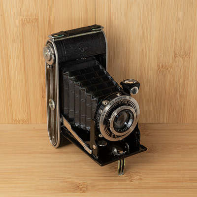 The image features an old-fashioned camera sitting on a wooden table. It is placed in the center of the scene, drawing attention to its vintage design. The camera appears to be black and silver, with a focus on the lens area.