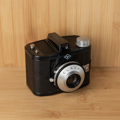 The image features a vintage black and silver camera sitting on top of a wooden table. The camera is an old-fashioned model, possibly a Polaroid or a Kodak, with a leather case surrounding it. The camera has a shiny metal lens, which adds to its antique charm.