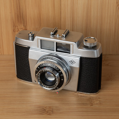 The image features an old-fashioned camera sitting on a wooden table. The camera is made of metal and has a leather case, giving it a vintage appearance. It appears to be an antique model, possibly from the 1950s or earlier.