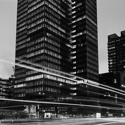 The image is a black and white photo of two tall buildings in the city. One building is located on the left side, while the other is situated towards the right. Both buildings are surrounded by traffic lights, with one set of traffic lights positioned near the center of the scene and another set closer to the right edge.