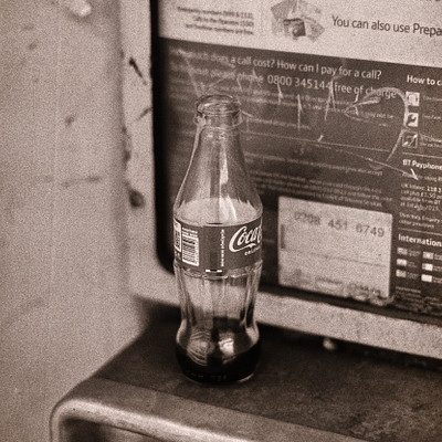 The image is a black and white photo of an old-fashioned telephone booth with a Coca Cola bottle sitting on top of it. The bottle appears to be empty, and the phone booth has a sign posted above it. The scene gives off a nostalgic vibe, reminiscent of times when public phones were more common.