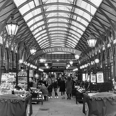 The image is a black and white photo of an indoor marketplace, likely in the Apple Market. There are several people walking around and shopping in the area. Some individuals can be seen carrying handbags as they browse through the market.