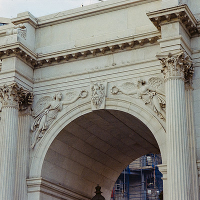 The image features a large, ornate stone archway with statues on its sides. There are several people standing under the arch, possibly admiring or taking pictures of the impressive structure. In addition to the main archway, there is another smaller arch visible in the background.