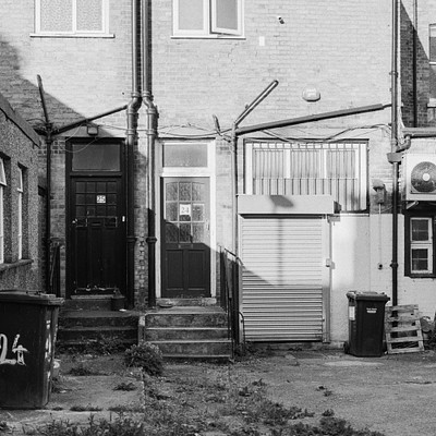 The image is a black and white photo of an alleyway between two brick buildings. There are several steps leading up to the door of one of the houses, which has a garage attached. A small shed can be seen in front of the house, and there's a trash can located near the bottom left corner of the image.