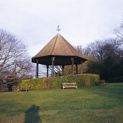 The image features a park with a gazebo, which is an open-sided structure with a roof. There are several benches placed around the gazebo, providing seating for visitors to relax and enjoy the surroundings. A person can be seen standing near one of the benches, possibly admiring the view or taking a break from walking.