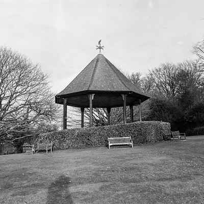 The image is a black and white photo of an outdoor park setting. In the center, there is a gazebo with a green roof, surrounded by trees. A bench can be seen in front of the gazebo, providing a place for visitors to sit and relax.