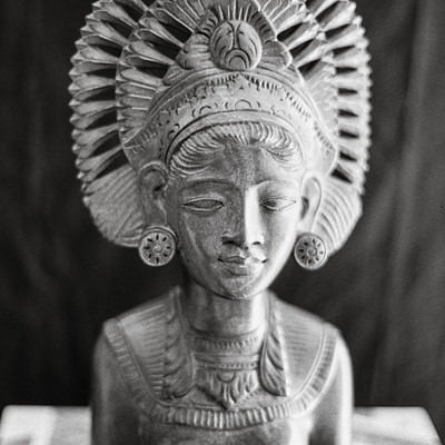 The image features a statue of a woman with a crown on her head, sitting in front of a black background. She is wearing earrings and has an intricate design on her headpiece. The statue appears to be made of metal or stone, giving it a unique and elegant appearance.