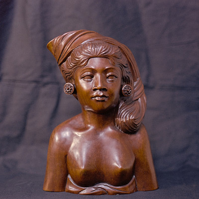 The image features a brown statue of a woman with a headband, possibly an Egyptian or African style. She is wearing a necklace and has a peaceful expression on her face. The statue appears to be made from wood and is placed in front of a black background.