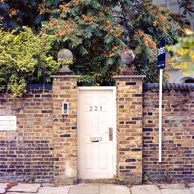 The image features a small white door with the number 213 on it, located in front of a brick wall. The door is situated between two pillars and appears to be an entrance or exit point for a building. A sign can also be seen near the door, possibly providing information about the location or directions.