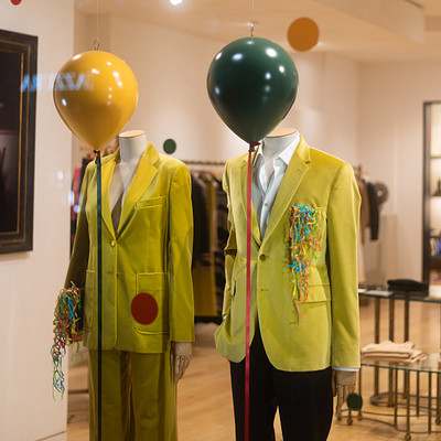 The image features a store display with two mannequins wearing yellow jackets. One of the mannequins has a green balloon on its head, while the other one has a yellow balloon. Both mannequins are dressed in suits and ties, creating an eye-catching presentation for potential customers.