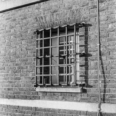 The image is a black and white photo of an old brick building with a window. The window has bars on it, giving the impression that it might be a jail or a similar type of structure. A power line can be seen running along the side of the building, adding to its aged appearance.