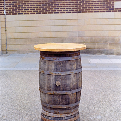 The image features a wooden barrel or crate, which has been repurposed as an outdoor table. It is placed on the ground in front of a brick wall and a building. The table appears to be made from a large wooden barrel, giving it a rustic and unique appearance.