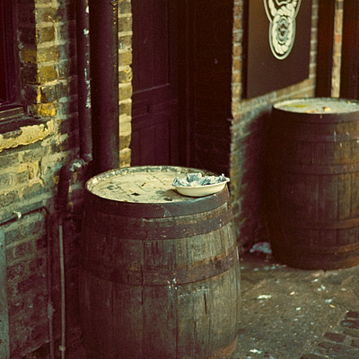 The image is a black and white photo of two wooden barrels sitting on the sidewalk in front of a building. One barrel is larger, occupying most of the space, while the other one is smaller. They are placed next to each other, with one slightly behind the other.