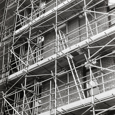 The image is a black and white photo of a building with scaffolding on it. There are multiple windows visible, some of which have bars covering them. The scaffolding appears to be made of metal and is attached to the side of the building.