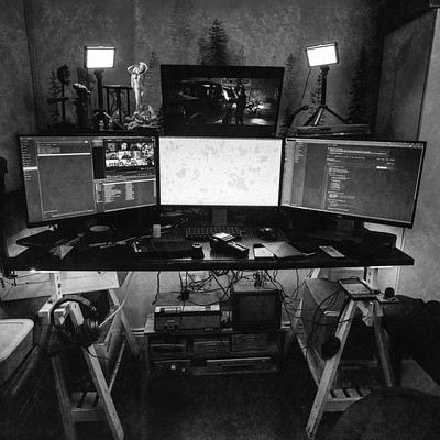 The image is a black and white photo of a computer desk with three monitors on it. There are two keyboards, one in front of each monitor, and two mice placed near the keyboards. A TV screen can also be seen on the left side of the desk.