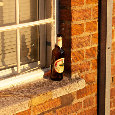A bottle of beer is sitting on a ledge outside, next to a brick wall. The bottle appears to be empty and has been left there for someone else to find or take.