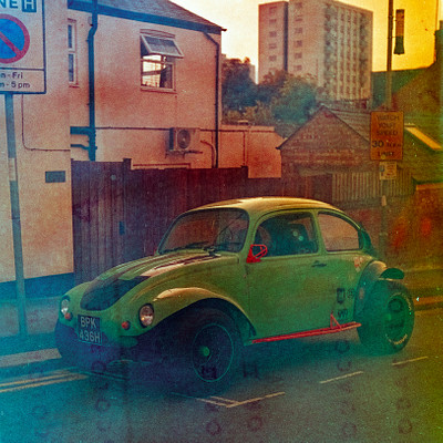 The image is a black and white photo of an old green Volkswagen Beetle car parked on the side of a street. It appears to be in front of a house, possibly near a fence or a building. There are two people visible in the scene, one standing closer to the left side of the car and another person further back on the right side.
