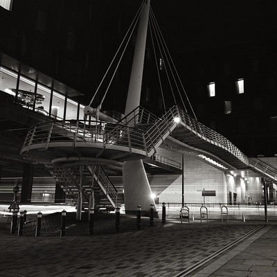 The image is a black and white photo of an outdoor area with a metal bridge crossing over a street. There are several benches placed around the scene, some near the bridge and others further away. A few people can be seen in the background, possibly walking or enjoying their time at this location.