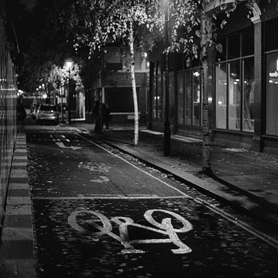 The image is a black and white photo of a city street at night. There are two bicycles parked on the sidewalk, one closer to the center of the scene and another further back. A car can be seen in the background, parked near the left edge of the image.