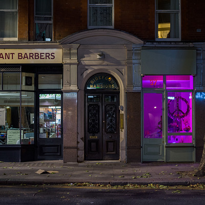 The image features a street corner with a barber shop called "The Pleasant Barbers" on the side of the road. The storefront is illuminated by purple lights, giving it an eye-catching appearance. There are several chairs visible in front of the building, possibly for customers to sit while getting their haircuts.