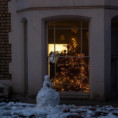The image features a snowman standing in front of a house, with its head sticking out from the doorway. The snowman is positioned next to a Christmas tree that has been decorated with lights and ornaments. The scene appears to be set on a street corner, as there are two potted plants visible in the foreground.