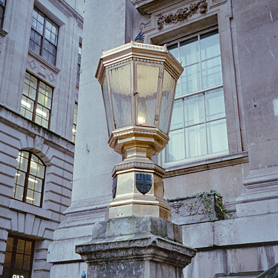 The image features a street light with a gold top, located in front of an old building. The lamp post is situated on the sidewalk and appears to be made of stone or concrete. There are several windows visible on the building, some of which have shutters.