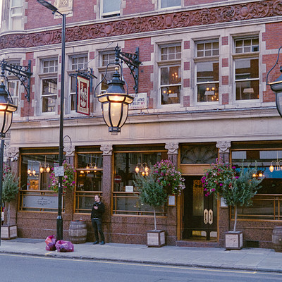 The image depicts a street scene with a large brick building featuring a restaurant. There are several potted plants placed around the area, adding greenery to the surroundings. A woman is standing outside of the building, possibly waiting for someone or just enjoying her time on the sidewalk.