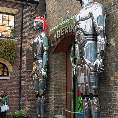 The image features a couple of large, silver robot statues standing on the sidewalk in front of a building. These robots are positioned next to each other and appear to be part of an art installation or decoration. In addition to the robots, there is a person visible near the left edge of the image, possibly admiring the sculptures or passing by.