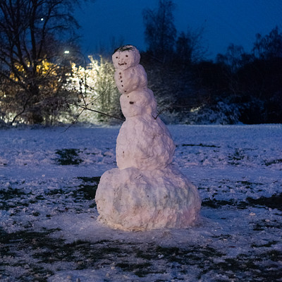 The image features a snowman standing in the middle of a field. It is made up of various snowballs and sticks, giving it a unique appearance. The snowman appears to be quite large, taking up most of the scene.