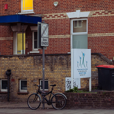 The image features a red brick building with a bicycle parked outside. There are two bikes visible in the scene, one near the center of the frame and another towards the left side. A person can be seen standing next to the bike on the right side of the building.