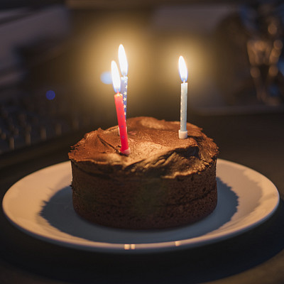 The image features a chocolate cake with two lit candles on top, placed on a white plate. The cake is sitting on a table next to a computer keyboard and mouse. There are also several books scattered around the scene, adding to the cozy atmosphere of the setting.