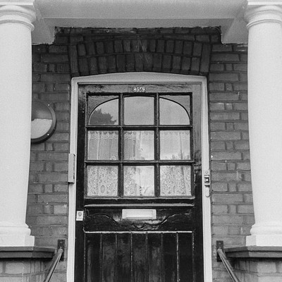 The image is a black and white photo of an old house with a brick exterior. A doorway is visible on the side of the building, featuring a window above it. There are two steps leading up to the door, and a railing can be seen in front of them.