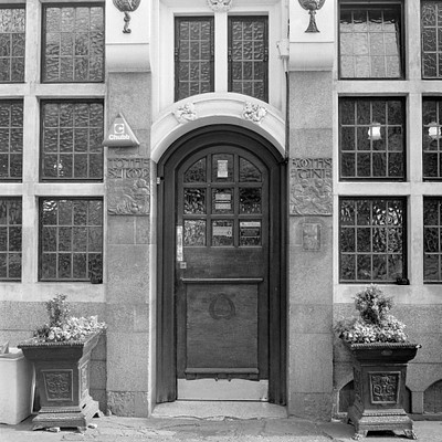 The image features a large, old building with an ornate doorway. The door is open and inviting, leading to the entrance of the building. Above the door, there are two potted plants adding a touch of greenery to the scene.