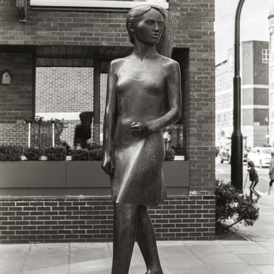 The image is a black and white photograph of a statue of a woman standing on a platform. She appears to be wearing a dress, and the statue has a unique design with wheels under her feet. The scene also includes several potted plants placed around the area, adding greenery to the environment.