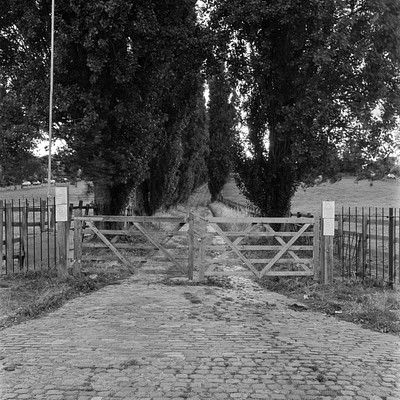 The image is a black and white photo of a dirt road surrounded by trees. There are two large wooden gates on the side of the road, one closer to the left side and the other nearer to the right side. A fence can be seen in front of the gates, adding an extra layer of security.