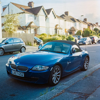 The image features a blue sports car parked on the side of a street. It is situated in front of several houses, with one house being particularly large and white. There are multiple cars parked along the street, including some smaller vehicles like a truck and a few other cars.