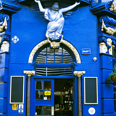 The image features a blue building with an ornate facade, including a statue of a woman hanging from the side. The building has a large arched doorway and several windows adorning its exterior. There are also potted plants placed around the area, adding to the overall ambiance of the scene.