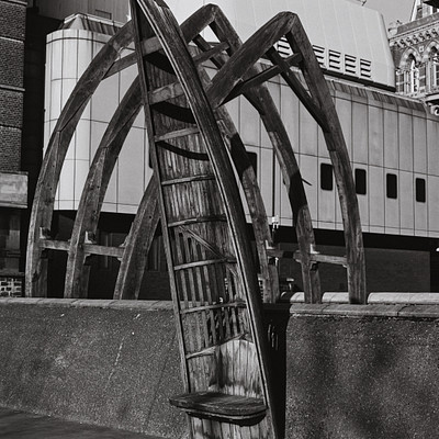The image is a black and white photo of an old wooden chair sitting on the sidewalk. It appears to be a unique piece of art or decoration, possibly made from an old ladder. The chair is positioned in front of a building with a clock tower visible in the background.