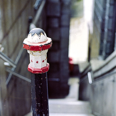 The image features a staircase with a black metal railing and a red pole. On the pole, there is a small statue of a man wearing a hat. The stairs are located in an urban setting, possibly near a subway station or another public transportation hub.