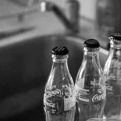 The image features a table with three bottles of Coca-Cola sitting on it. Each bottle has a black cap, and they are all placed next to each other in a row. The bottles appear to be empty, as there is no liquid visible inside them.