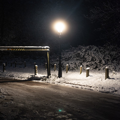 The image depicts a snowy street at night with a lamppost shining brightly. The light illuminates the area, creating a serene atmosphere. There are several benches placed along the sidewalk, providing seating for pedestrians.